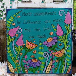 May include: A square, hand-painted artwork with a teal background and a dark blue border. The artwork features colorful flowers and the text: "Never underestimate the difference you make and the lives you touch."