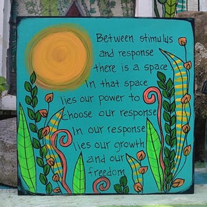 May include: A square, hand-painted teal sign with the text "Between stimulus and response there is a space..." in black. The sign features a yellow sun and colorful botanical illustrations in green, orange, and pink.