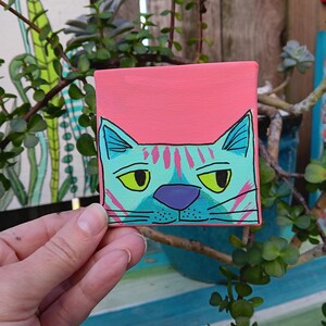 Peut inclure: Petite peinture sur toile carrée représentant un visage de chat stylisé. Le chat est turquoise avec des rayures roses, des yeux jaunes et un nez violet, sur fond rose corail. L'œuvre est tenue par une main, avec de la verdure en arrière-plan.