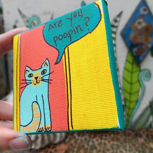 May include: A small square canvas painting featuring a light blue cat with orange stripes on its tail. The cat is positioned against a red and yellow background. A teal speech bubble above the cat contains the black text, Are you poopin?