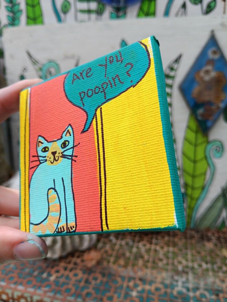 May include: A small square canvas painting featuring a light blue cat with orange stripes on its tail. The cat is positioned against a red and yellow background. A teal speech bubble above the cat contains the black text, Are you poopin?