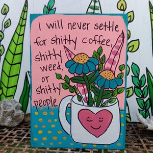 May include: A rectangular wooden sign with a pink background and a blue base featuring yellow dots. It displays a white mug with a pink heart face containing blue flowers and green leaves. The text reads: I will never settle for shitty coffee, shitty weed, or shitty people.