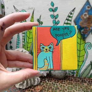 May include: A small square canvas painting featuring a light blue cat with orange stripes on its tail. The cat is positioned against a red and yellow background. A teal speech bubble above the cat contains the black text, Are you poopin?