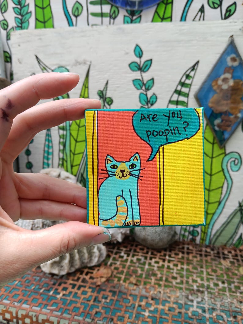 May include: A small square canvas painting featuring a light blue cat with orange stripes on its tail. The cat is positioned against a red and yellow background. A teal speech bubble above the cat contains the black text, Are you poopin?