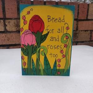Puede incluir: Un letrero de madera pintado de amarillo con flores verdes y rosas y el texto "Bread for all and roses too".