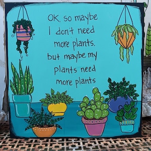 Peut inclure: Une plaque en bois carrée avec un fond turquoise vif présentant des illustrations peintes à la main de diverses plantes en pot et suspendues. Le texte indique : OK, so maybe I don't need more plants, but maybe my plants need more plants.