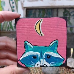 May include: A small square canvas painting featuring a teal raccoon face with white markings and black eyes, set against a solid pink background. Above the raccoon is a yellow crescent moon. The painting has a thick black border around the edges.