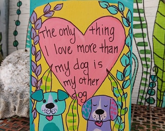 May include: A rectangular wooden sign with a yellow background and a large pink heart in the center. The text reads, The only thing I love more than my dog is my other dog. Two cartoon dogs are painted below the heart, one teal and one purple.