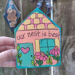 May include: A hand-painted wooden house-shaped sign with the text "our nest is best". The sign features a teal door with a pink heart, pink flowers, and a yellow and pink roof. The sign is held in front of a wooden fence.