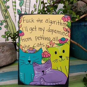 May include: A hand-painted wooden sign with a yellow background and black border. The sign features three cartoon cats in teal, purple, and lime green. The text reads "Fuck the algorithm, I'll get my dopamine from petting all the cats."