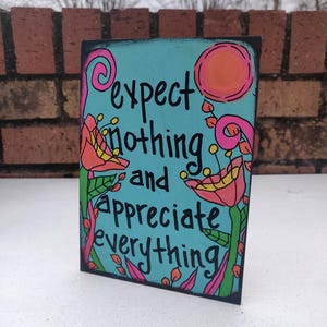 May include: A rectangular wooden sign with a teal background and black border. The sign features the words "expect nothing and appreciate everything" in black script. Floral and sun illustrations in pink, orange, and green decorate the sign.
