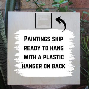 以下が含まれることがあります： 「Paintings ship ready to hang with a plastic hanger on back」と黒文字で書かれた白い木製の看板です。