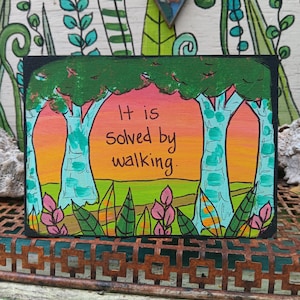 May include: A rectangular wooden sign featuring a hand-painted landscape with green trees, a vibrant orange and pink sunset sky, and pink foliage. The text "It is solved by walking." is written in black ink in the center of the scene.