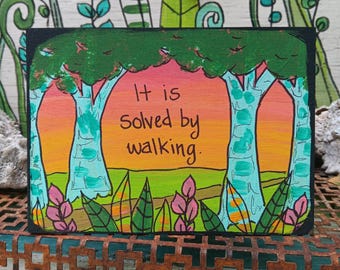 May include: A rectangular wooden sign featuring a hand-painted landscape with green trees, a vibrant orange and pink sunset sky, and pink foliage. The text "It is solved by walking." is written in black ink in the center of the scene.