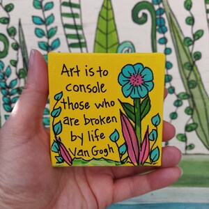 Puede incluir: Un pequeño lienzo cuadrado amarillo con la cita "El arte es para consolar a los que están rotos por la vida - Van Gogh". Una flor turquesa y hojas rosas están pintadas en el lienzo.