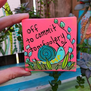 May include: A small, square, pink canvas with the words "off to commit tomfoolery" in black. A cartoon snail with a blue shell and yellow body is depicted, surrounded by green leaves and grass. The artwork is held by a hand.