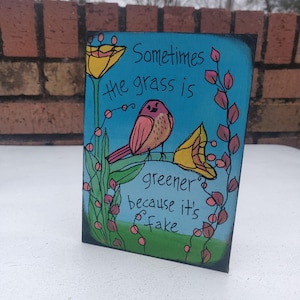 May include: A blue and green painted wooden sign with a pink bird and yellow flowers. The sign reads "Sometimes the grass is greener because it's fake."