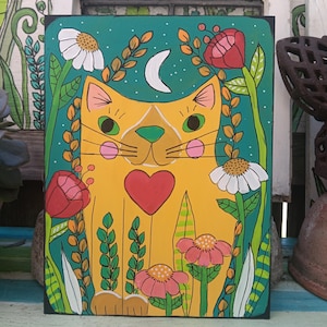 May include: A whimsical painting of a yellow cat with a red heart, surrounded by flowers and foliage. The cat has green eyes and pink cheeks. A white crescent moon is in the teal background. The artwork is on a black rectangular wooden panel.