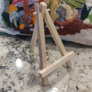 May include: A small, unfinished wooden easel with a three-legged design. The easel is made of light-colored wood and is designed to hold a small canvas or artwork. The background features a colorful, embroidered pillow with mushroom and floral designs.