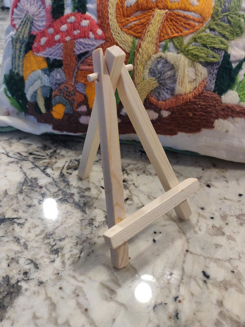 May include: A small, unfinished wooden easel with a three-legged design. The easel is made of light-colored wood and is designed to hold a small canvas or artwork. The background features a colorful, embroidered pillow with mushroom and floral designs.