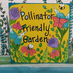 Puede incluir: Un letrero de madera pintado a mano con un fondo amarillo y las palabras "Pollinator Friendly Garden" en negro. El letrero está decorado con ilustraciones coloridas de flores, abejas y mariposas, con un borde turquesa.