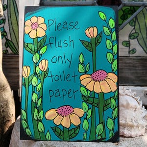 May include: A rectangular sign with a teal background and black border. The sign features hand-painted yellow and pink flowers with green stems and leaves. The text "Please flush only toilet paper" is written in black.