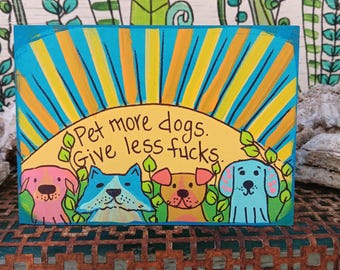 May include: A rectangular wooden sign featuring a hand-painted scene with four colorful dogs in front of a yellow sunburst background. The text "Pet more dogs. Give less fucks." is written in black ink across the center of the yellow sun.