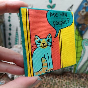 May include: A small square canvas painting featuring a turquoise cat with a yellow nose and tail sitting in a doorway. The background consists of a red panel and yellow borders. A turquoise speech bubble contains the black text Are you poopin?