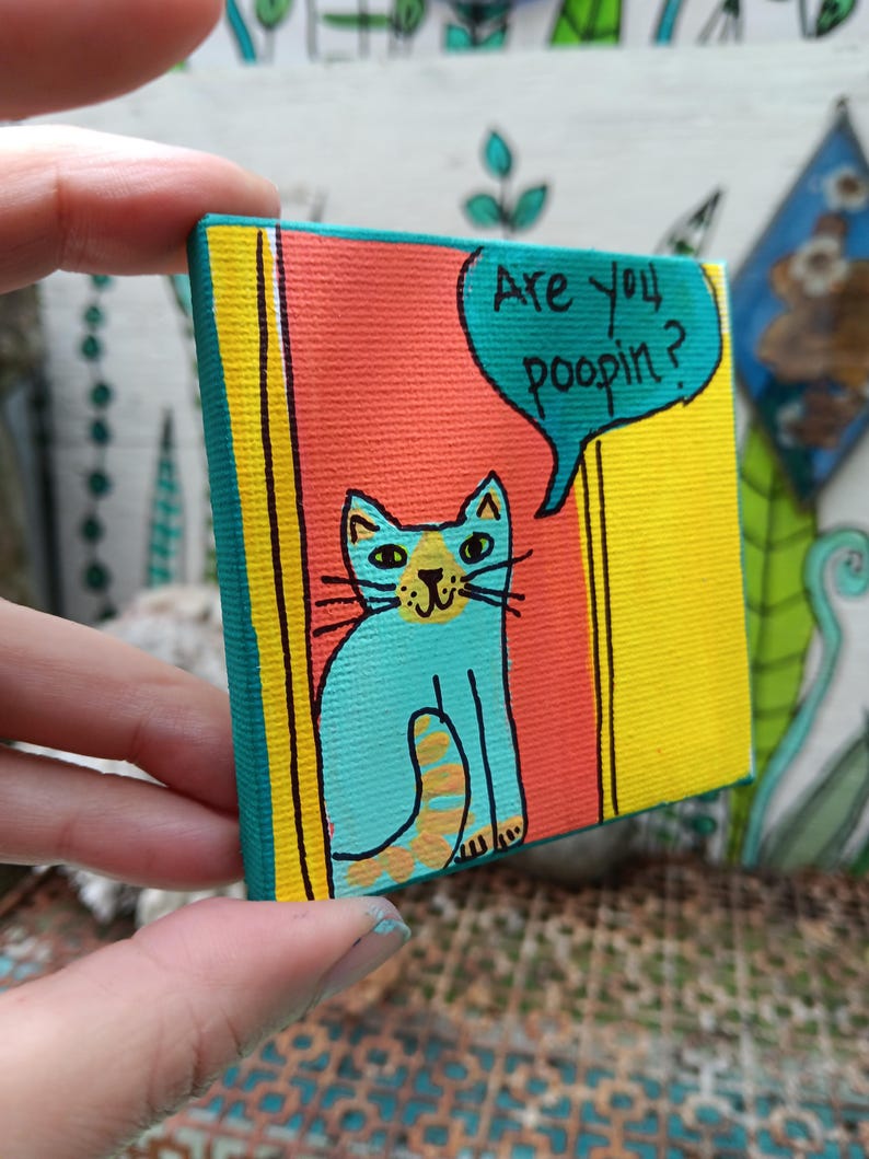 May include: A small square canvas painting featuring a turquoise cat with a yellow nose and tail sitting in a doorway. The background consists of a red panel and yellow borders. A turquoise speech bubble contains the black text Are you poopin?
