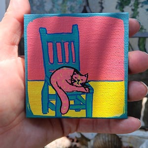 May include: A small square canvas painting featuring a pink cat resting on a teal chair. The background is divided into a horizontal pink upper section and a yellow lower section. The cat is outlined in black with simple facial features.