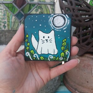 May include: A small, square painting featuring a white cat under a full moon in a starry night sky. The cat is surrounded by green foliage. The painting has a black border and is held in a hand.