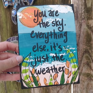 Könnte beinhalten: Ein rechteckiges, handgemaltes Kunstwerk mit schwarzem Rand. Das Kunstwerk zeigt einen blauen Himmel, eine orangefarbene Sonne und eine Meereslandschaft mit den Worten "You are the sky. Everything else, it's just the weather."