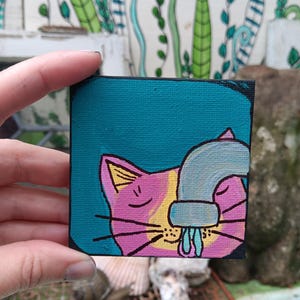 May include: A small square canvas painting featuring a pink and yellow cat with closed eyes. A light blue faucet is positioned over the cat's nose, with a small stream of blue water dripping down. The background is a solid dark teal color.