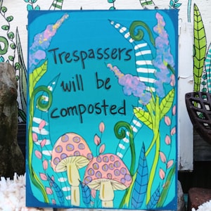 May include: A square, hand-painted sign with a turquoise background and the text "Trespassers will be composted." The sign features two pink and purple spotted mushrooms, surrounded by green and blue foliage.
