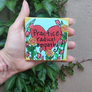 May include: A small square canvas with a yellow border and a light blue background. The artwork features a red heart with the words "Practice radical empathy" in black. The heart is surrounded by green leaves, pink buds, and yellow roses.