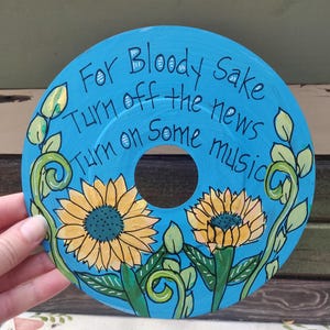 May include: A hand-painted, repurposed vinyl record with a bright blue background. The record features two yellow sunflowers with green leaves and vines. The text "For Bloody Sake Turn off the news Turn on Some music" is written in black.