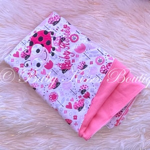 Op de afbeelding: Een roze en witte babydeken met een lieveheersbeestje ontwerp en de tekst "Baby Kisses".