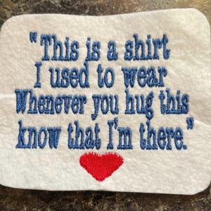 May include: White embroidered patch with blue text that reads "This is a shirt I used to wear Whenever you hug this know that I'm there." A red heart is embroidered below the text. The patch is rectangular with rounded corners.