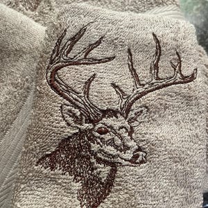 May include: Tan bath towel with a detailed embroidered image of a deer's head and antlers in shades of brown. The towel has a textured surface and a folded edge.