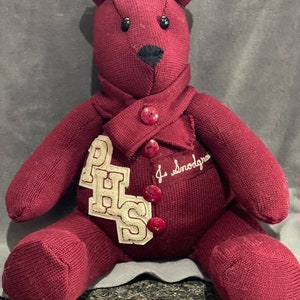 May include: A maroon teddy bear wearing a maroon sweater with white lettering that says "PHS" and a white collar with two red buttons. The sweater has a white tag that says "J. Snodgrass".