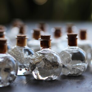 May include: A group of small, clear glass bottles with cork tops filled with silver flakes. The bottles are arranged in a row on a dark surface.