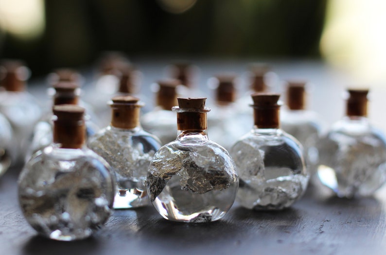 May include: A group of small, clear glass bottles with cork tops filled with silver flakes. The bottles are arranged in a row on a dark surface.