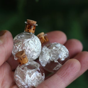May include: Three small, round glass bottles with cork stoppers. The bottles are filled with a clear liquid and silver flakes.