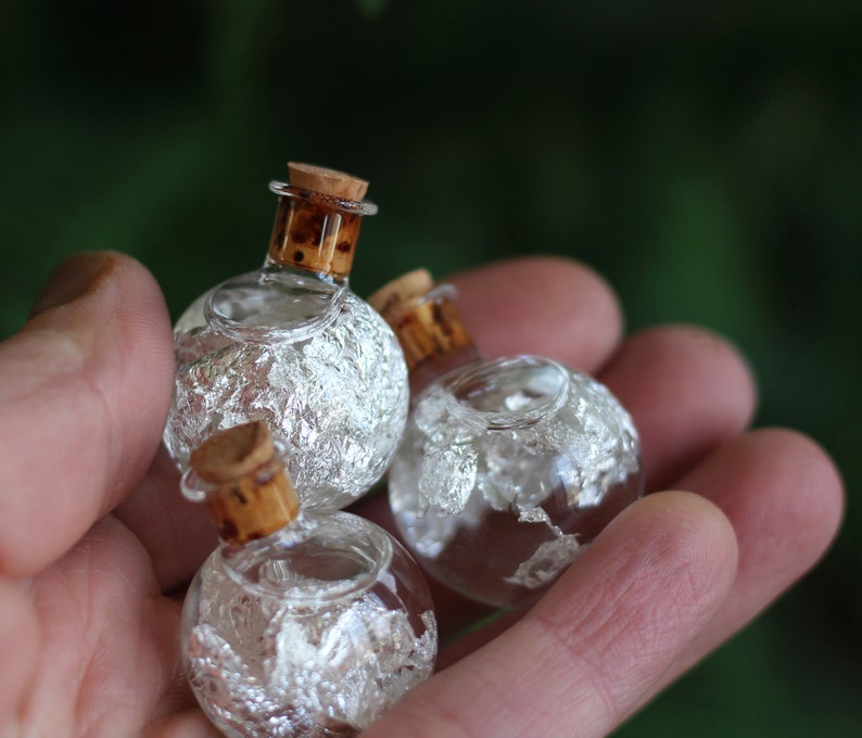 May include: Three small, round glass bottles with cork stoppers. The bottles are filled with a clear liquid and silver flakes.