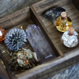 May include: A wooden box with a divided interior, containing various items. The left side features a small orange crystal, a dried flower, and a large clear quartz crystal. The right side contains two small glass bottles with cork stoppers, one filled with a golden liquid and the other with a silver liquid.