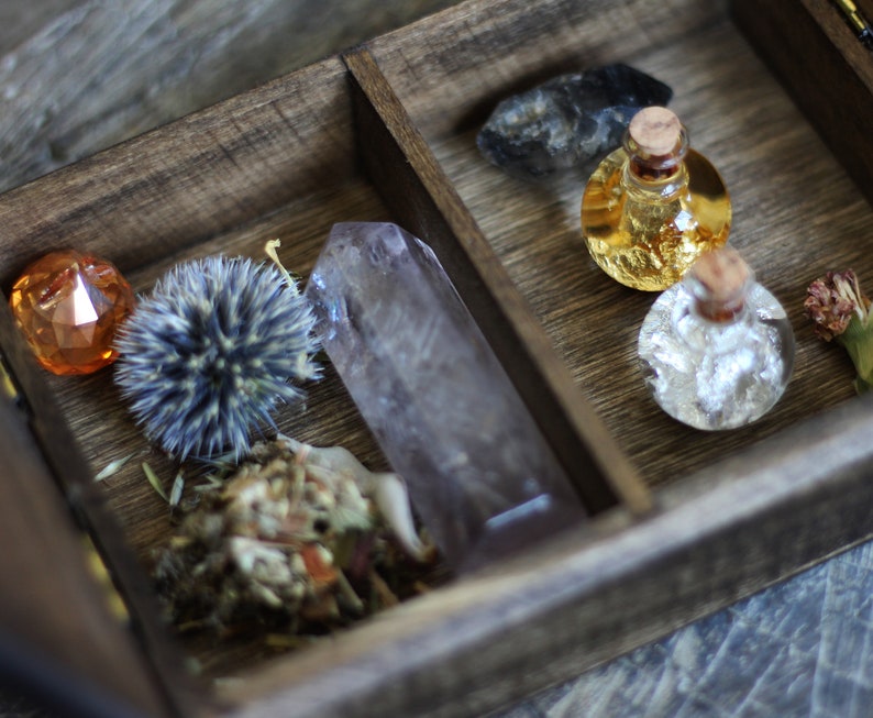 May include: A wooden box with a divided interior, containing various items. The left side features a small orange crystal, a dried flower, and a large clear quartz crystal. The right side contains two small glass bottles with cork stoppers, one filled with a golden liquid and the other with a silver liquid.