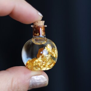 May include: A small, clear glass bottle with a cork stopper. The bottle is filled with a clear liquid and gold flakes.