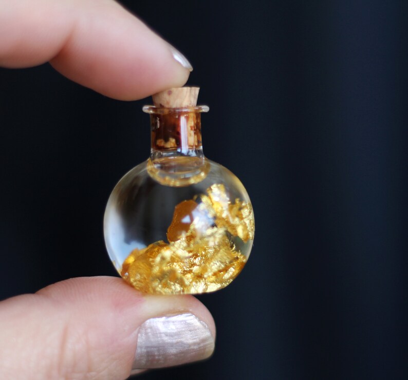 May include: A small, clear glass bottle with a cork stopper. The bottle is filled with a clear liquid and gold flakes.