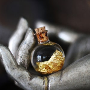 May include: A small glass bottle with a cork stopper, filled with gold flakes, resting in the palm of a hand-shaped sculpture.