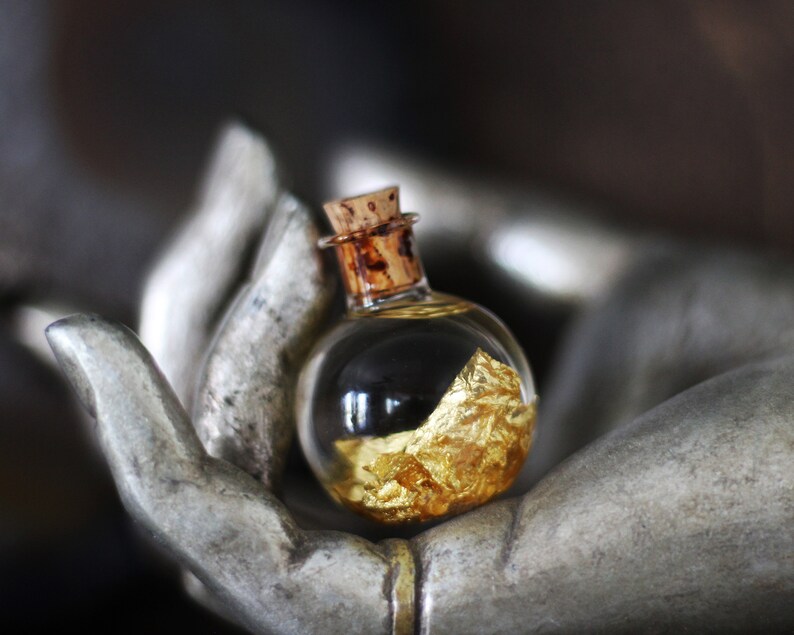 May include: A small glass bottle with a cork stopper, filled with gold flakes, resting in the palm of a hand-shaped sculpture.